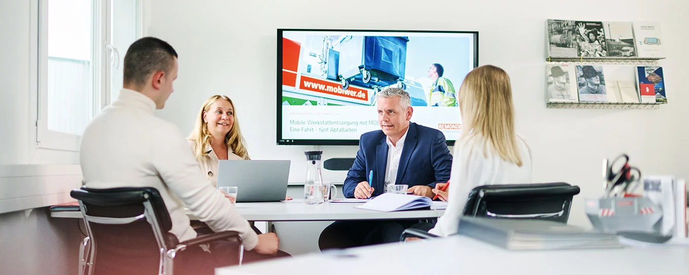 Vier Mitarbeiter von REMONDIS Industrie Service in einer Besprechung an einem Tisch sitzend; im Hintergrund ist ein Fernseher mit einem MOBIWER-Fahrzeug darauf zu sehen
